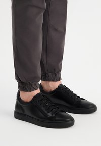 Czarne skórzane trampki z zaokrąglonymi czubkami, wiązanym designem i bocznym zamkiem błyskawicznym. Nosi się je z szarymi spodami tapered, które mają elastyczne mankiety.