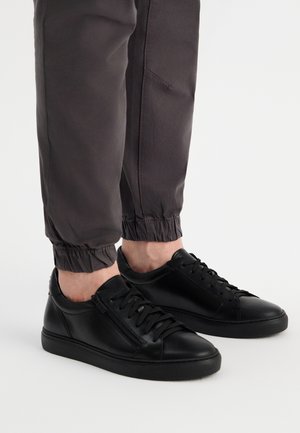 Czarne skórzane trampki z zaokrąglonymi czubkami, wiązanym designem i bocznym zamkiem błyskawicznym. Nosi się je z szarymi spodami tapered, które mają elastyczne mankiety.