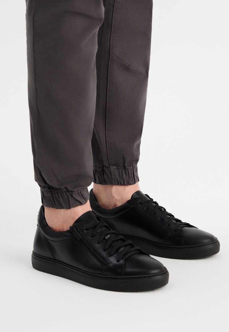 Czarne skórzane trampki z zaokrąglonymi czubkami, wiązanym designem i bocznym zamkiem błyskawicznym. Nosi się je z szarymi spodami tapered, które mają elastyczne mankiety.