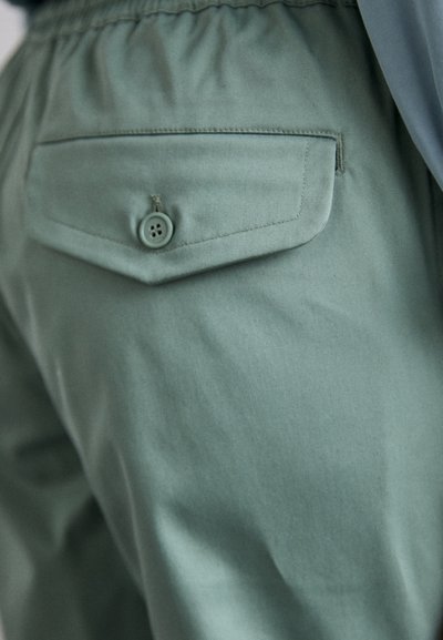Zelené kalhoty s knoflíkovou zadní kapsou, elastickým pasem a hladkou texturou, zdůrazňující minimalistický design.