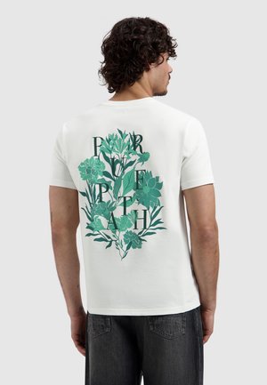 Man die een wit T-shirt draagt met een groen bloemmotief en de letters "PORTRAIT" op de achterkant, gecombineerd met donkergrijze jeans.
