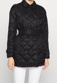 Czarna pikowana kurtka dwurzędowa z paskiem w talii, z zapięciem na guziki na mankietach i przeszyciem w diamentowy wzór, noszona z białymi spodniami.