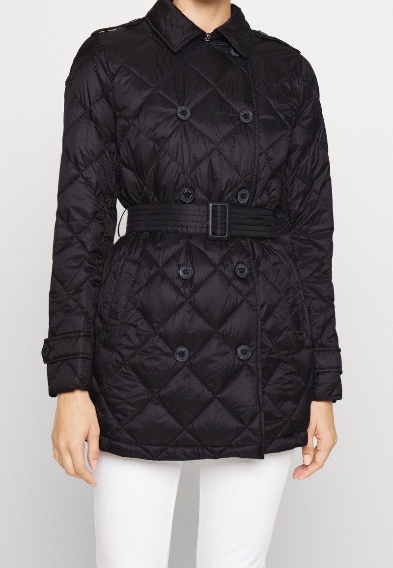 Czarna pikowana kurtka dwurzędowa z paskiem w talii, z zapięciem na guziki na mankietach i przeszyciem w diamentowy wzór, noszona z białymi spodniami.