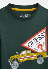Maglietta verde a maniche corte con grafica di un camion giallo e un triangolo rosso con il marchio "GUESS". Materiale in cotone, scollo rotondo.