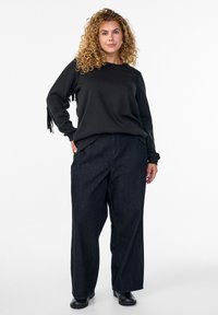 Čierna mikina s dlhým rukávom a bočnými strapcami, skombinovaná so voľnými tmavými džínsami. Čierne členkové topánky dotvárajú outfit.