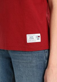 Camiseta de algodón roja con una etiqueta blanca con acentos azules y rojos que dice "NUEVA YORK, TOMMY JEANS." Manga corta, corte clásico.