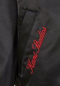 Černá látka s vertikálním štepířovým kapsou a červeným vyšitým textem "Kani Studios" vedle kapsy.