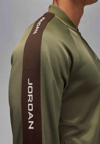 Olivgrön vindjacka med hög krage och en brun sidrand med "JORDAN" i vitt. Mjuk textur, skräddarsydd passform.