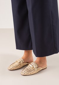 Mules de oro tejido con punta redonda, hebilla decorativa y suela de madera plana. Llevados con pantalones de pierna ancha en azul marino.