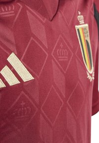 Maillot de football rouge avec un motif en losanges, arborant un logo Adidas doré et un blason belge. Tissu doux avec un design de col classique.