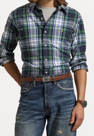 Man draagt een groen-wit-blauw geruit overhemd met opgestropte mouwen, bruine leren riem, blauwe spijkerbroek en een bruin horloge aan de linkerpols.