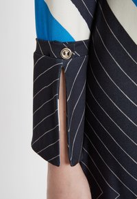 Ärmel eines marineblauen Kleidungsstücks mit weißen diagonalen Streifen. Besitzt einen Schlitz und einen dekorativen Knopf. Das Material wirkt glatt und leicht.