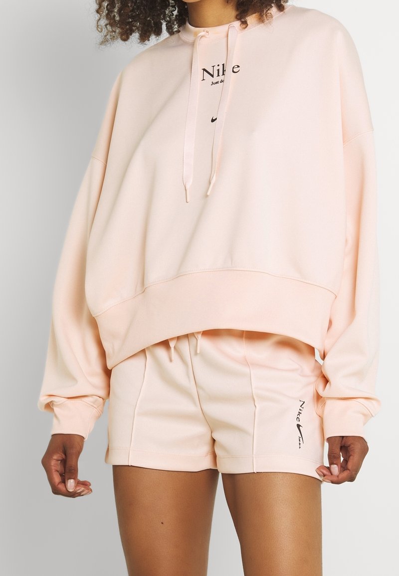 Personne portant un sweat à capuche oversize Nike rose clair et un short assorti avec des logos Nike noirs sur la poitrine et la cuisse gauche.
