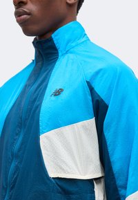 Jeune personne portant une veste zippée New Balance bleue et blanche avec un col montant, montrée de l’épaule au menton sur un fond uni.