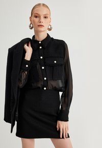 Schwarzes Zweiteiler-Outfit mit einer strukturierten, kurz geschnittenen Jacke und einer transparenten Bluse mit Knopfdetails. Der Rock ist figurbetont und strukturiert.