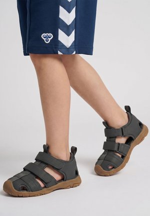 Barn iført navy shorts og grå lukkede sandaler med brune såler, stående på en ensfarvet lys baggrund.