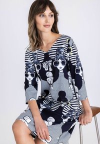 Vrouw met schouderlang haar, gekleed in een zwart-witte jurk met een herhaald beeld van vrouwen met een zonnebril, zittend op een kruk.