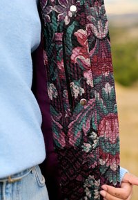 Gesteppte Jacke mit floralem Muster in lila-, grün- und burgunderfarbenen Tönen. Verfügt über einen vorderen Druckknopfverschluss und eine Seitentasche mit glatter Textur.