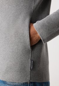Hånden satt inn i høyre lomme på grå strikkegenser merket Marc O’Polo, båret over blå denimjeans.