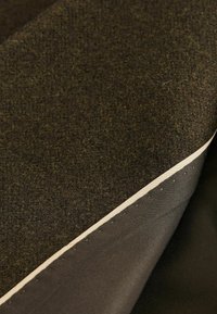 Dvě vrstvy látky: na vrchu je texturovaná tmavě hnědá vlna a pod ní hladký tmavý materiál s viditelným krémovým okrajem.