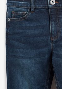 Jeans in denim blu scuro con cuciture arancioni, tasca frontale con rivetto, passante per cintura e chiusura con bottone metallico visibili.