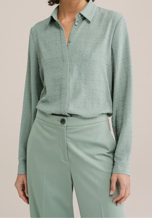 Vrouw die een lichtgroene, gestructureerde blouse met lange mouwen en knopen draagt, ingestopt in bijpassende, op maat gemaakte, hoog uitgesneden broek.