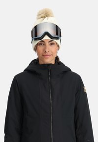 Schwarze Skijacke mit Kapuze, ausgestattet mit strukturierten Einsätzen und Reißverschluss. Getragen mit einer cremefarbenen Mütze und dunklen Skibrillen.
