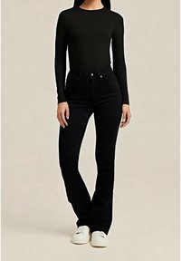 Femme portant un haut noir à manches longues, un pantalon noir taille haute et des baskets blanches, debout devant un fond uni.