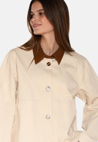 Beige jas met een bruine leren kraag, voorzien van zilveren knopen en zijzakken. Gemaakt van een gestructureerde stof.