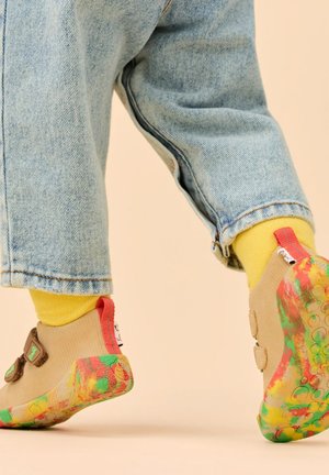 Kind trägt beige Schuhe mit mehrfarbiger Sohle, gelbe Socken und hochgekrempelte hellblaue Jeans vor hellem Hintergrund.