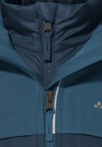 Veste isolée bleue avec col montant, fermeture éclair centrale et un tissu texturé. Présente des panneaux contrastants et un logo doré.