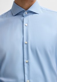 Jasnoniebieska koszula zapinana na guziki, wykonana z teksturowanej tkaniny z subtelnym diagonalnym wzorem. Posiada tradycyjny kołnierzyk i białe guziki.