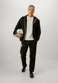 Vīrietis melnā sporta apģērbā ar dzelteniem akcentiem tur futbolu, stāvot uz bezrakstu gaiši pelēka fona.