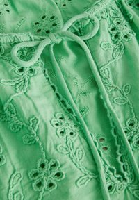 Tissu vert avec broderie florale et un nœud noué fait de bandes de tissu assorties, mettant en valeur des coutures décoratives texturées et des détails découpés.