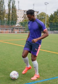 Fußballspieler in lila und pinkem Puma-Outfit mit kurzen Ärmeln und Shorts, der einen weißen Fußball auf einem grünen Feld tritt.