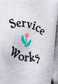 Γκρι ύφασμα με μαύρο κείμενο «Service Works» καμπυλωμένο γύρω από μια απλή γραφική απεικόνιση λουλουδιού με ροζ κύκλο και δύο πράσινα φύλλα.