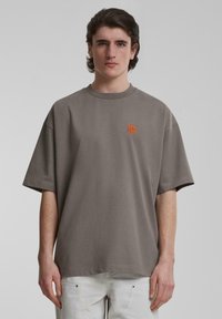 Camiseta de algodón gris de gran tamaño con cuello redondo y mangas cortas. Presenta un pequeño logo bordado naranja en el pecho. Textura lisa.