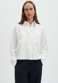 Camicia bianca con collo e bottoni, in tessuto testurizzato, con due tasche anteriori e polsini con bottoni, abbinata a pantaloni scuri.