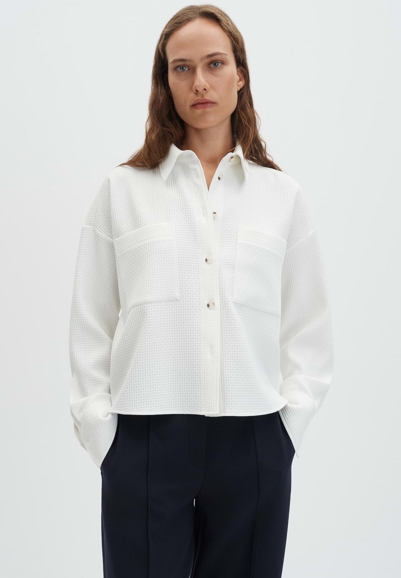 Camicia bianca con collo e bottoni, in tessuto testurizzato, con due tasche anteriori e polsini con bottoni, abbinata a pantaloni scuri.