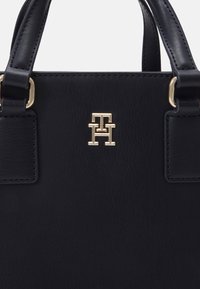 Svart lærveske med gullfarget hardware, med en fremtredende logo i sentrum og strukturerte håndtak. Glatt tekstur.