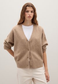 Beige gestrickte Strickjacke mit V-Ausschnitt, Knöpfen vorne und gerippten Bündchen. Kombiniert mit einem weißen Hemd und hellen Hosen.