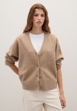 Beige knitted cardigan with a v-neck, buttons down the front, and ribbed cuffs. Paired with a white shirt and light-colored pants.
