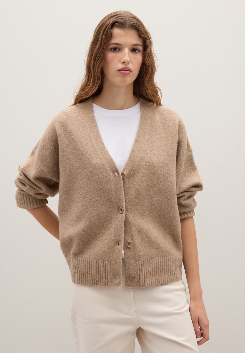 Beige gestrickte Strickjacke mit V-Ausschnitt, Knöpfen vorne und gerippten Bündchen. Kombiniert mit einem weißen Hemd und hellen Hosen.