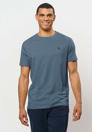 Uomo sorridente che indossa una maglietta blu tinta unita a maniche corte e pantaloni scuri, in piedi davanti a uno sfondo chiaro.