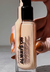 Fond de teint Always On de Smashbox dans un flacon en verre transparent avec une pompe noire, présentant une teinte beige liquide débordant de la buse.