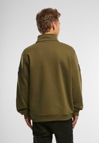 Felpa verde oliva con colletto alto, vestibilità comoda e maniche lunghe. Presenta toppe testurizzate su entrambe le maniche e un orlo a coste.