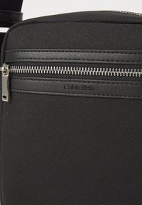 Sac bandoulière en tissu noir avec surface texturée, comprenant une fermeture zippée en haut et une petite plaque logo. Matériel de fermeture éclair en métal visible.