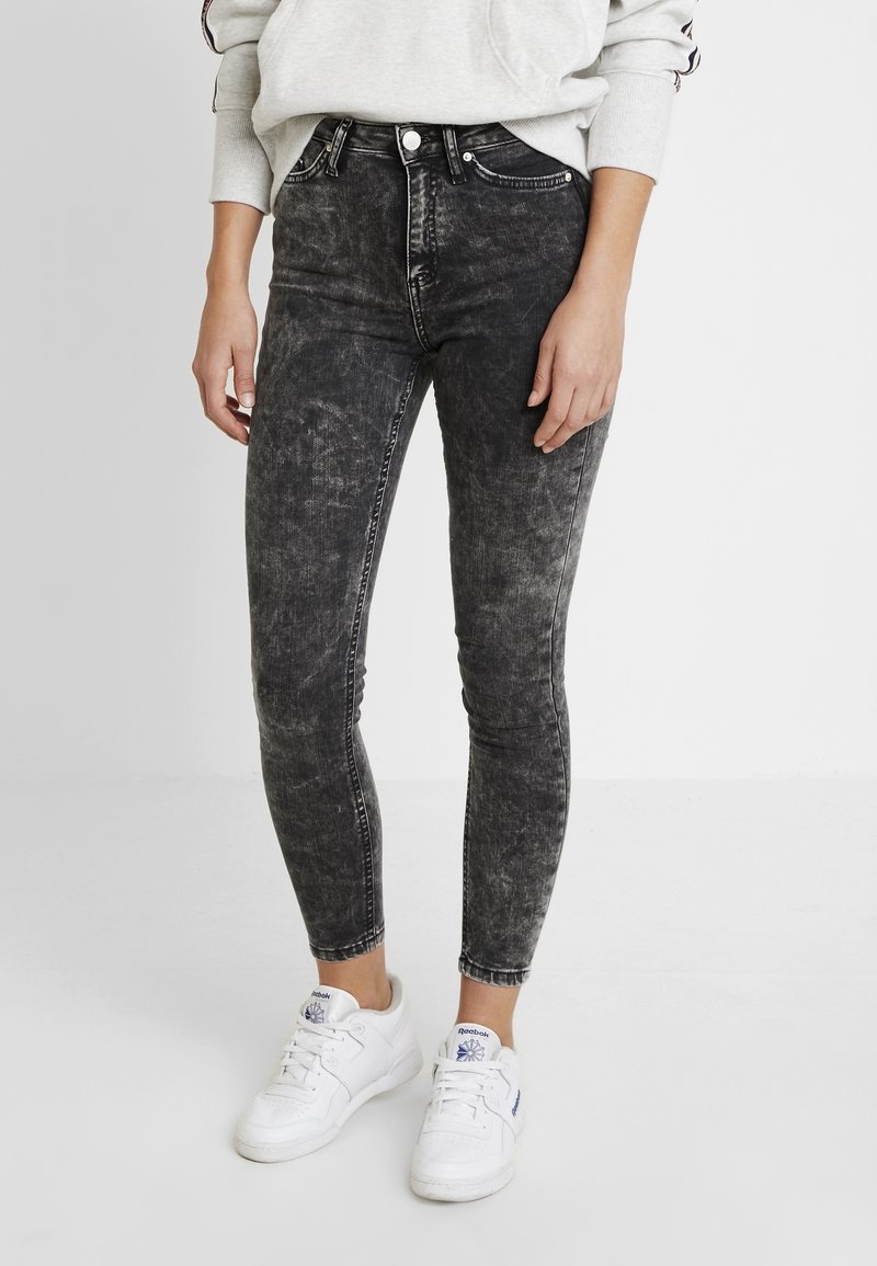 Personne portant un jean skinny noir délavé à l'acide, des baskets blanches et un sweat-shirt gris clair, debout devant un fond uni.