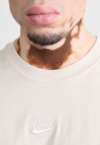 Ανοιχτό μπεζ βαμβακερό T-shirt με στρογγυλή λαιμόκοψη και κεντητό λευκό λογότυπο Nike στο στήθος. Ορατό δερματικό υπερχρωματισμό.