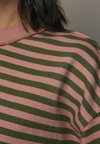 Primo piano di una persona che indossa un maglione a righe orizzontali rosa e verdi con colletto a costine e cuciture grezze.